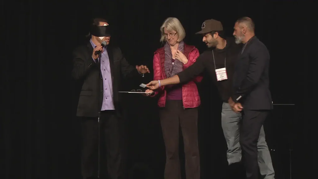 Randy Charach, blindfolded, and doing magic and mentalism act with audience participants.